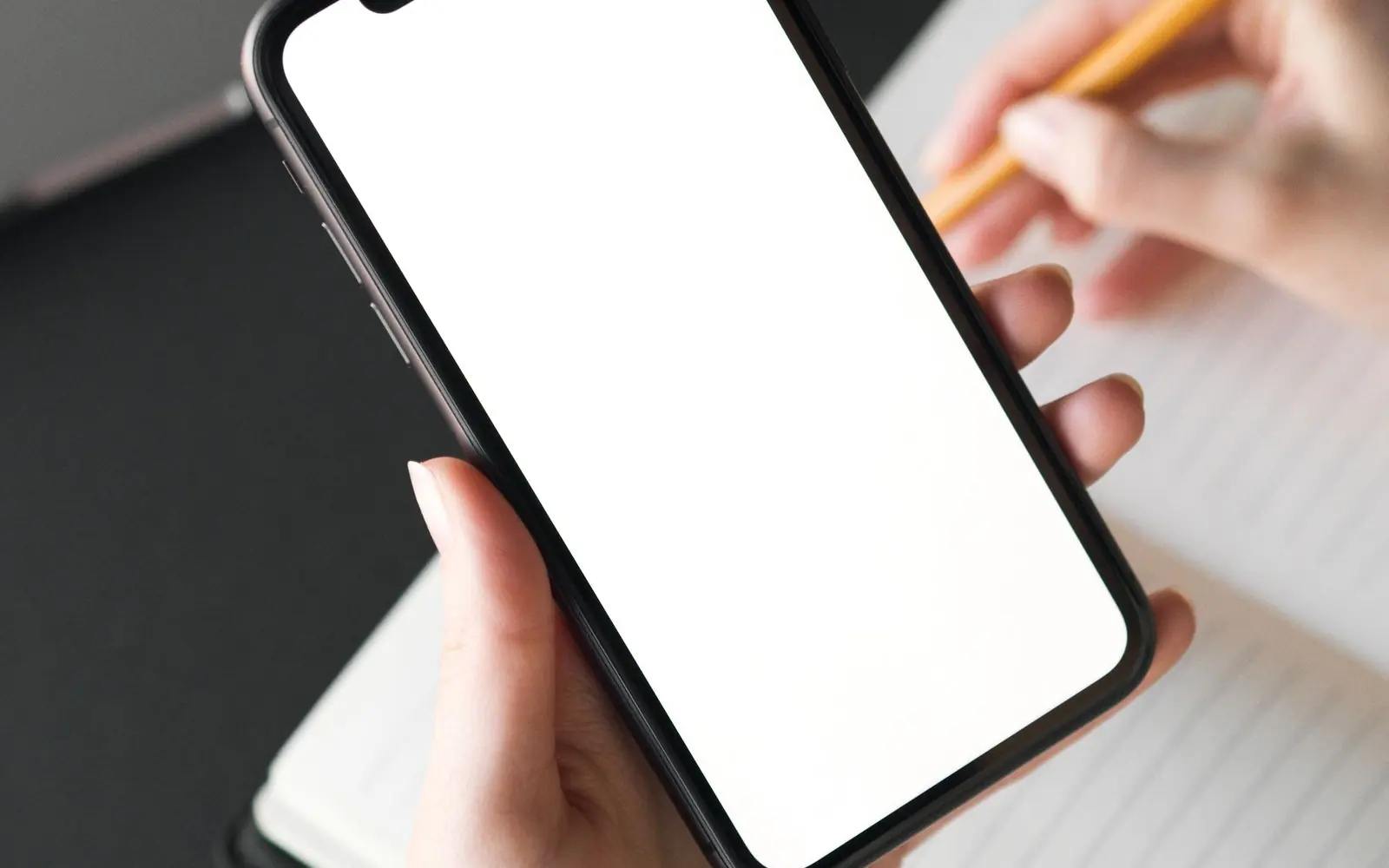 Mano sosteniendo un teléfono móvil con la pantalla en blanco mientras se toman notas en un cuaderno